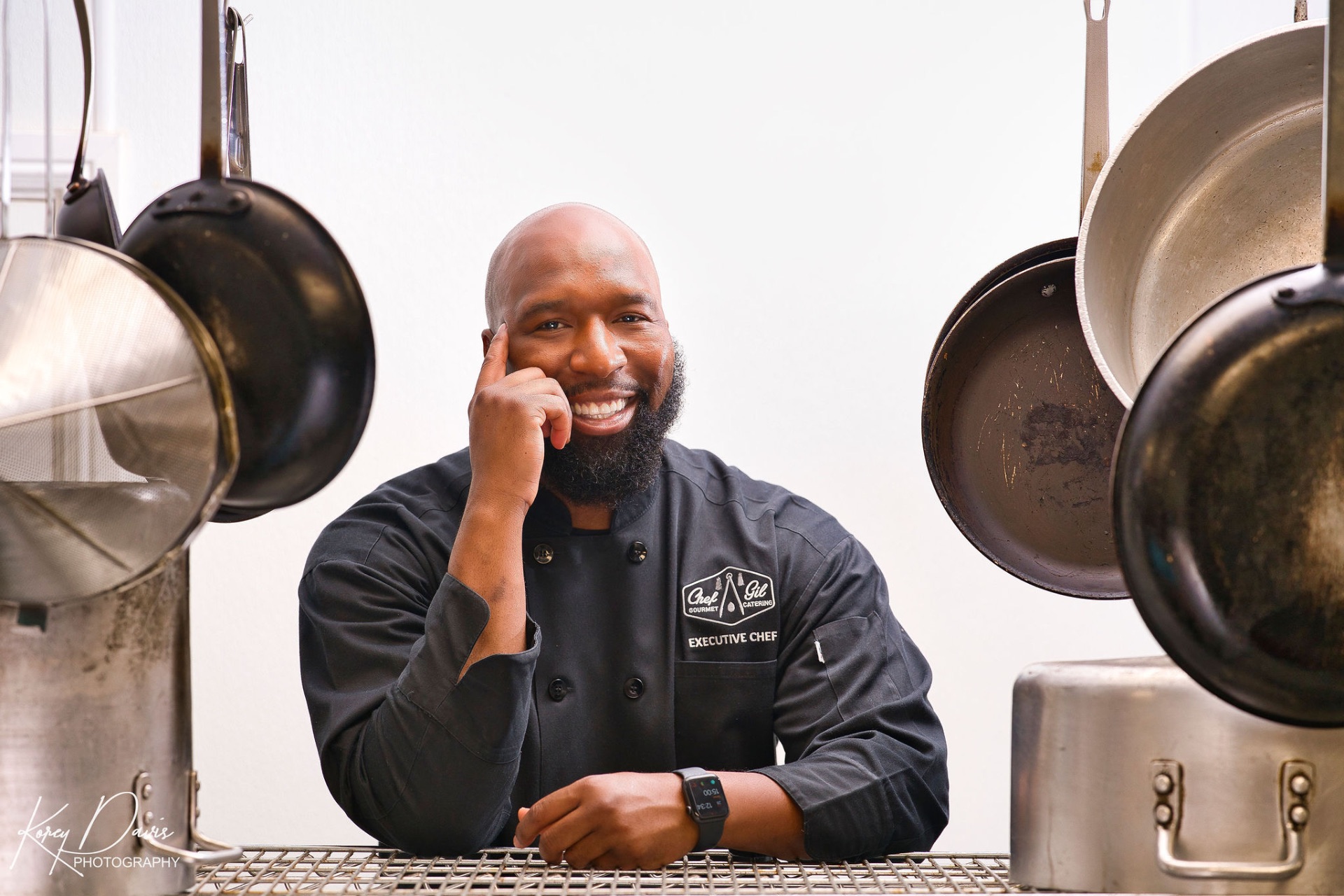 Chef Gilfert Hunt smiling in his black executive chef coat, framed by kitchen cookware